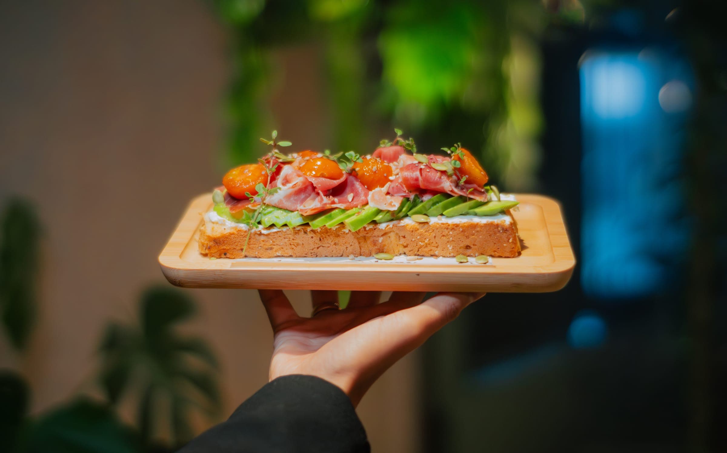 Una mano sostiene una tabla con tostada de palta, jamón y tomates en BAW Coffee